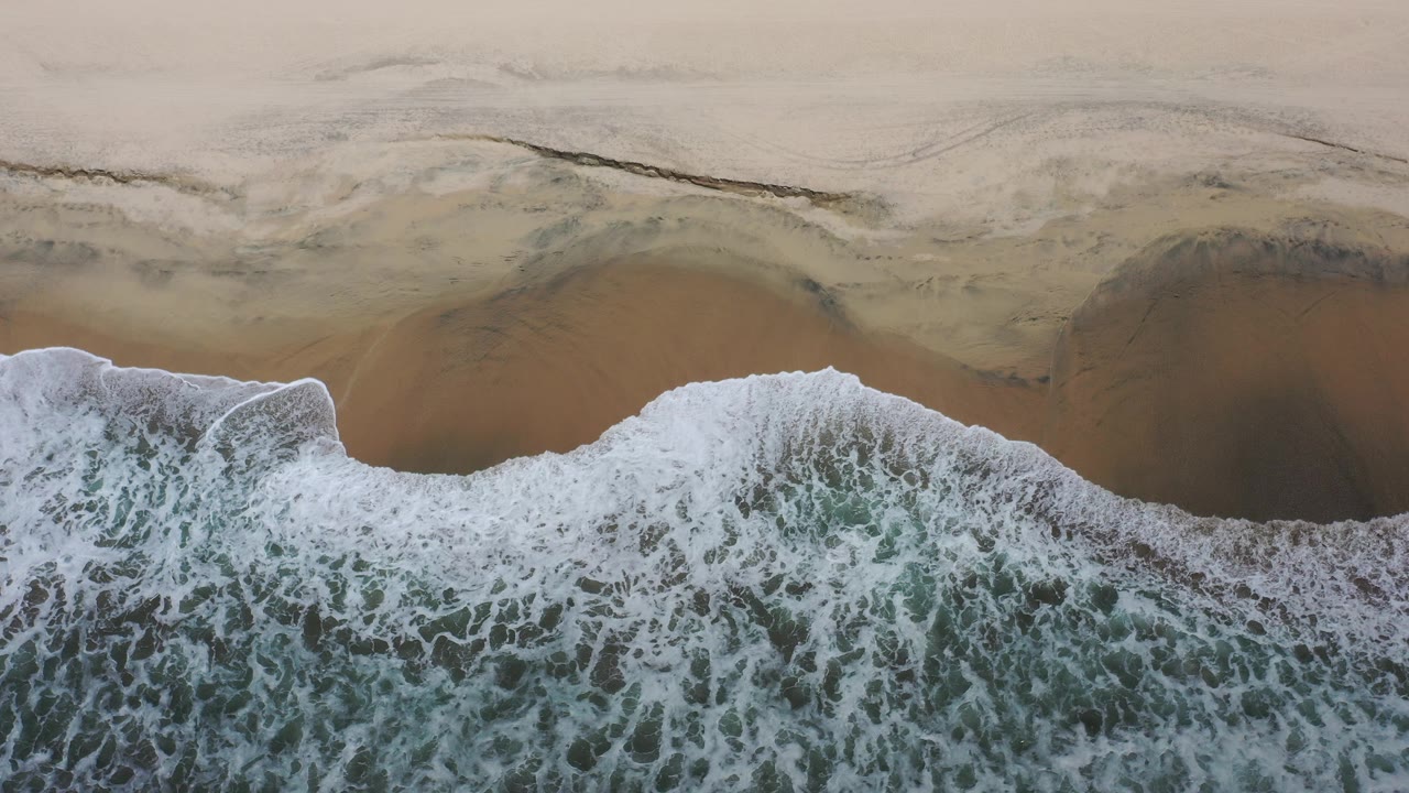 Overhead view in Los Cabos, Mexico, with a wide band of white surf meeting golden sand and no visible people.