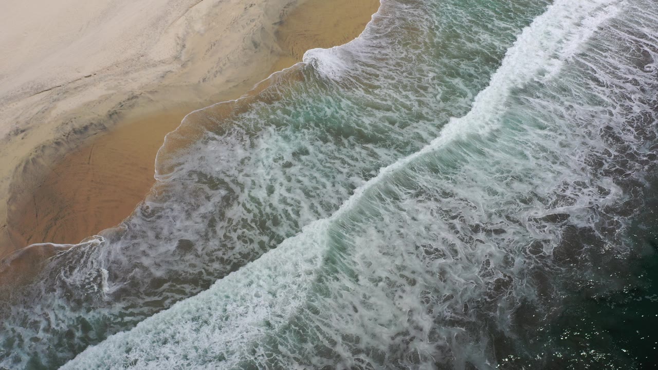 Overhead view of Los Cabos surf washing diagonally onto tan sand, with white foam spread across green-blue water and no visible people.