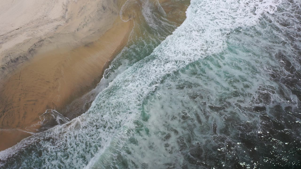 Overhead view of white surf pushing across tan beach and dark teal water in Los Cabos, Mexico, with no visible people.