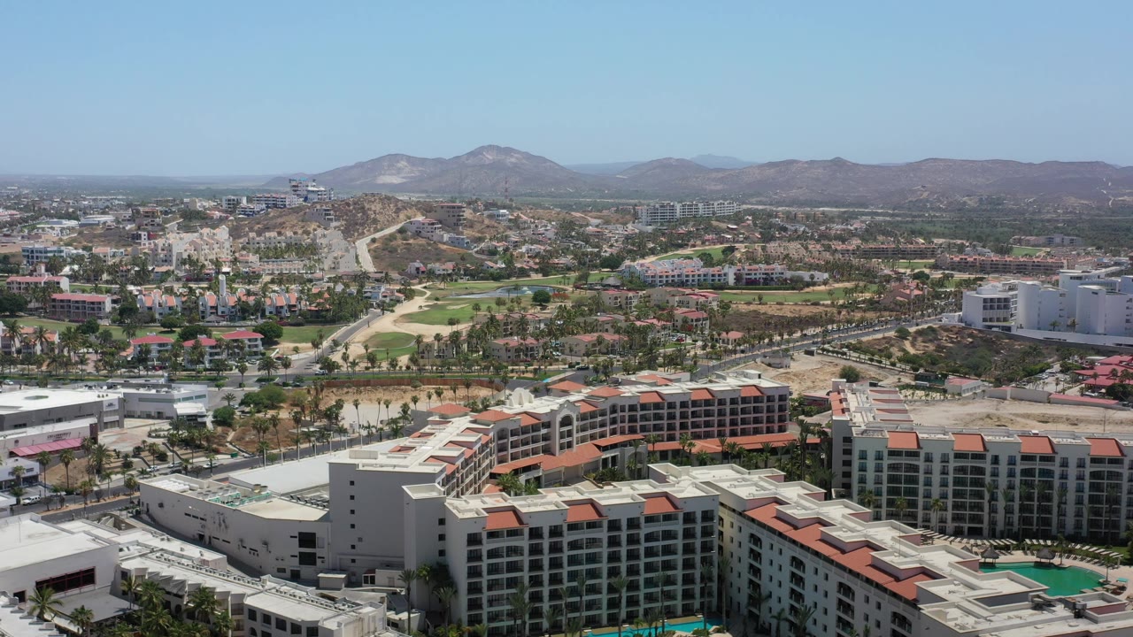 Opening high view over Los Cabos with white resort buildings and terracotta roofs in the foreground, roads and green fairways beyond, dry hills, distant mountains, and no visible people.
