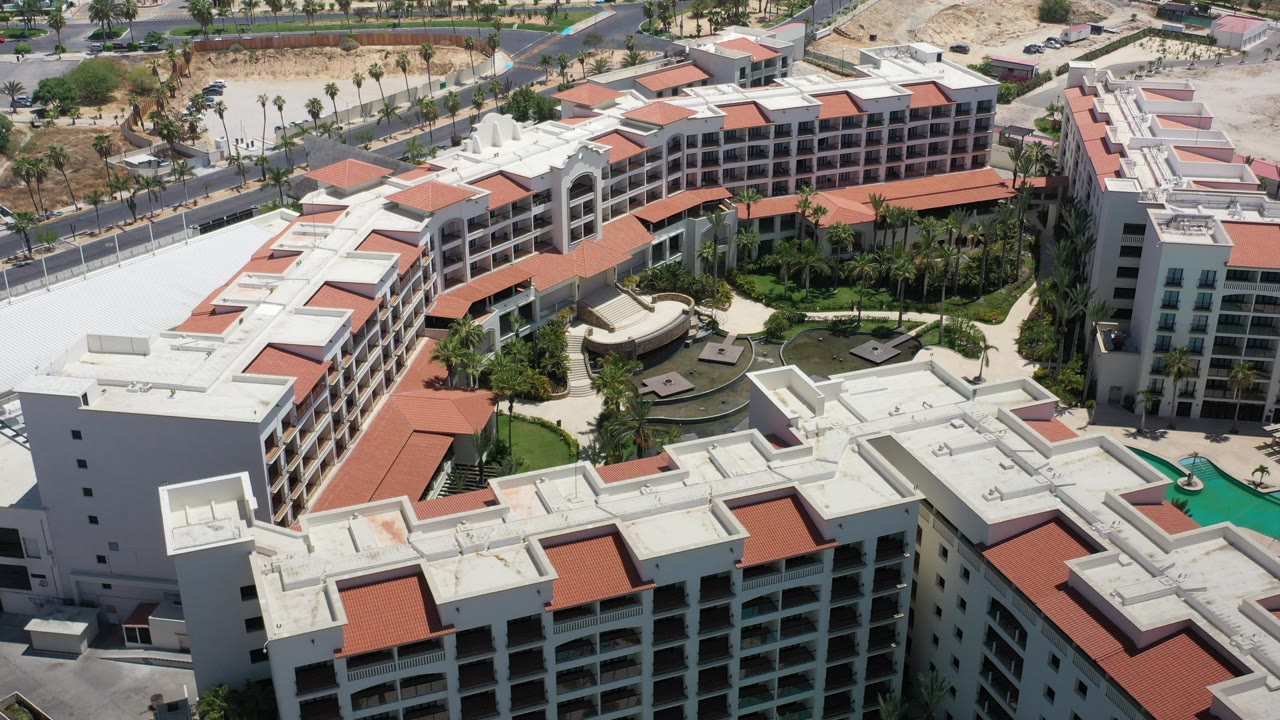 A high aerial frame over a Los Cabos hotel complex shows white multi-story buildings, red tile roof sections, inner courtyards, turquoise pool water, surrounding streets, and no visible people.