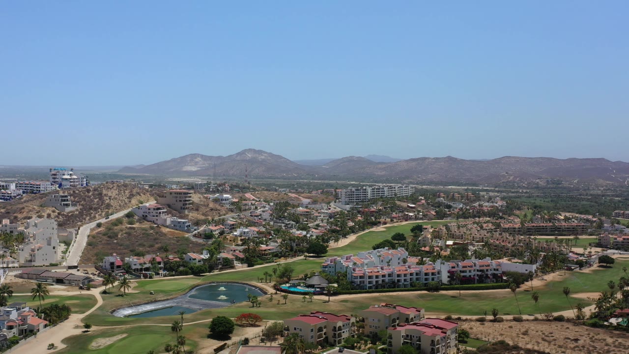 Los Cabos aerial view with pale roads climbing a dry hill at left, fairways and a pond below, clustered homes in the midground, mountains on the horizon, and no visible people.