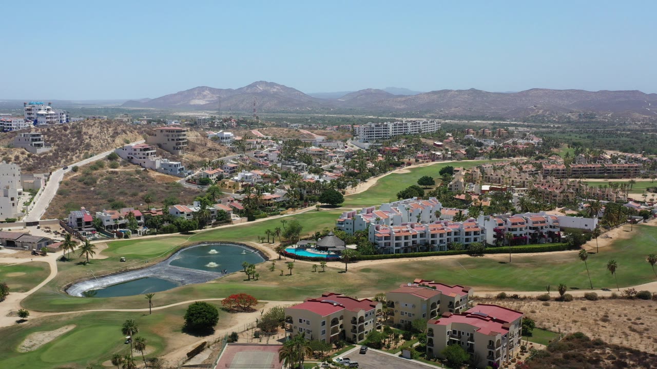 Los Cabos aerial frame showing green golf fairways, two water features, red-roofed resort buildings, dry hills, and distant mountains with no visible people.
