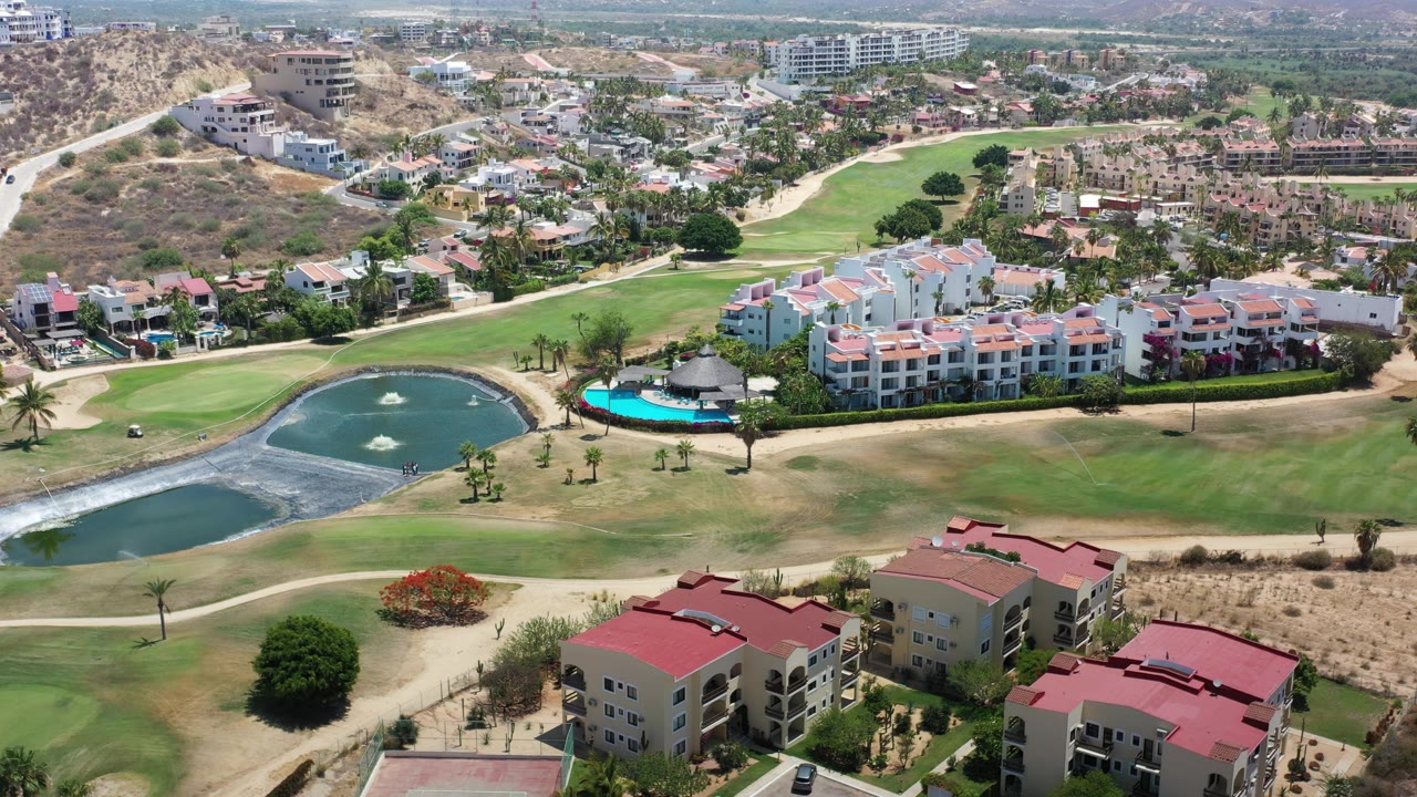 Drone aerial in Los Cabos, Mexico with two ponds, fountain spray, green fairways, a turquoise swimming pool beside low-rise buildings, foreground rooftops, and no visible people.
