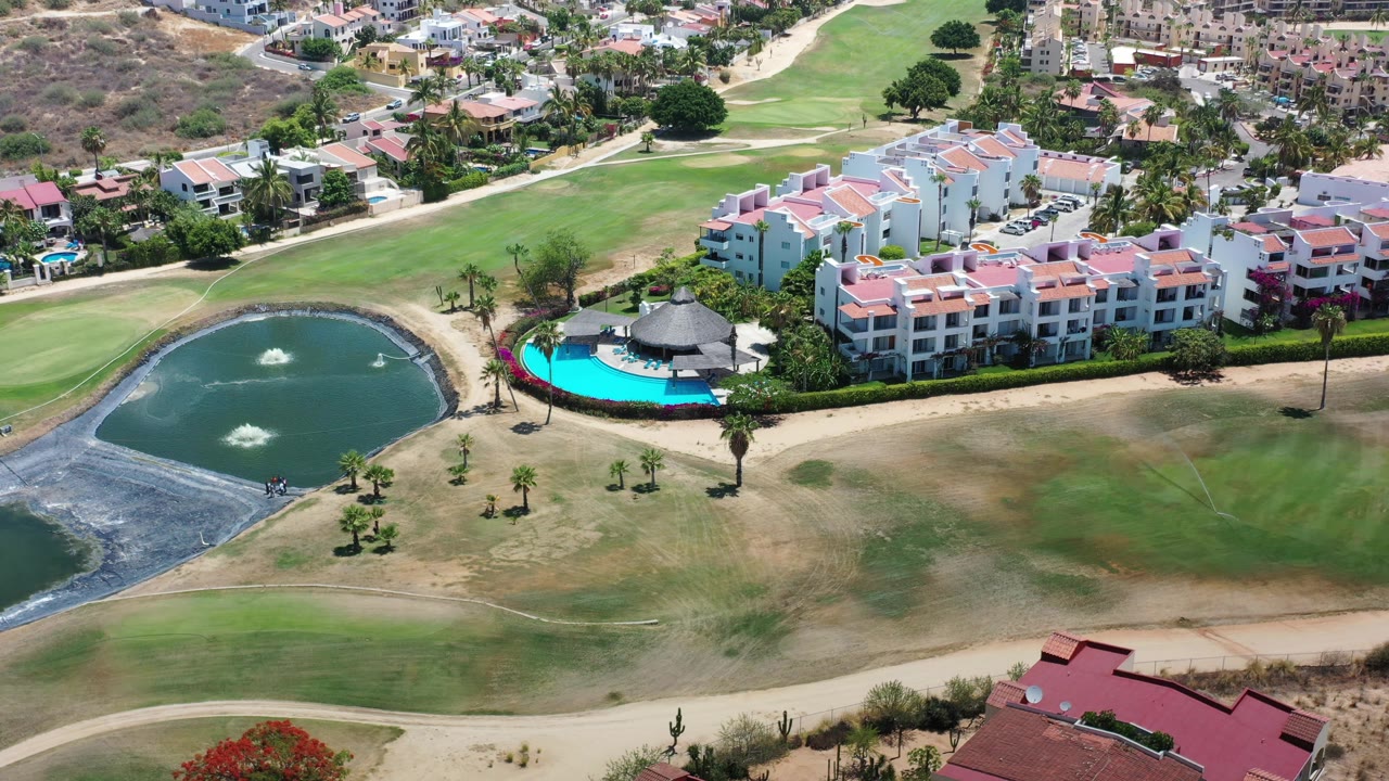 Aerial view in Los Cabos with the pond and fountains left of frame, a bright blue pool beside multi-story villas, golf-course grass, access roads, and no visible people.