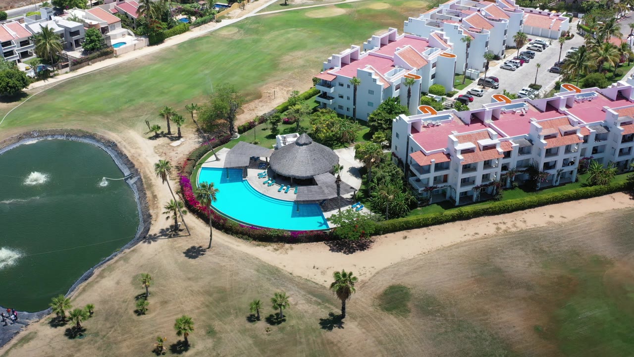 Aerial frame over Los Cabos residential buildings with the bright pool near center, the pond fountain at left, palms along the path, parked cars near the upper right, and tiny distant people near the lower-left pond edge.