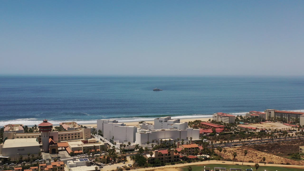 Opening aerial frame over Los Cabos, Mexico with blue ocean, a sandy beach, beachfront buildings, red tile roofs, dry ground, and no visible people.