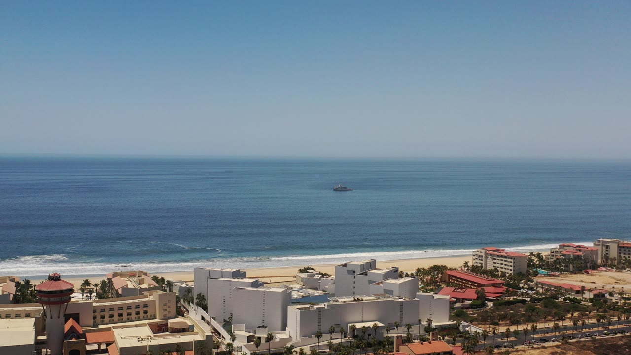 A wide Los Cabos coastal view shows resort buildings at the bottom of frame, pale beach and surf beyond them, and open blue ocean under clear sky with no visible people.