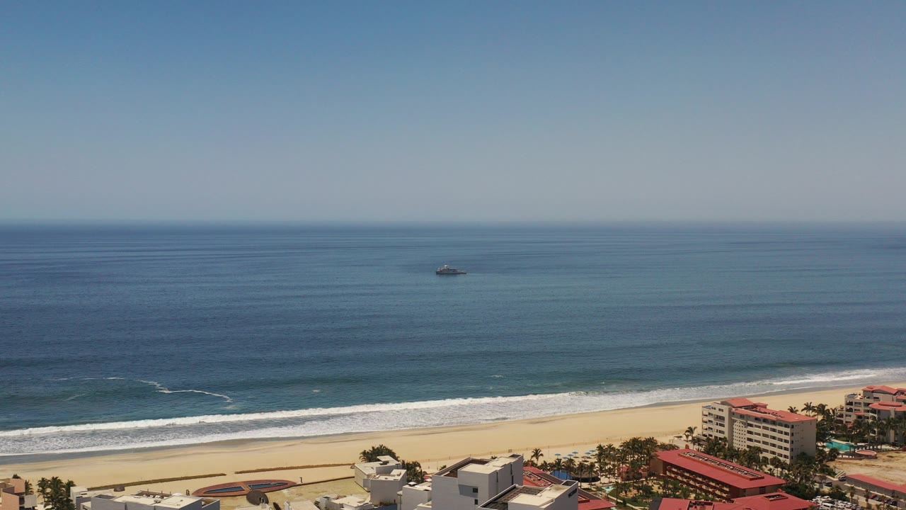 Wide aerial view in Los Cabos, Mexico, showing blue ocean, a distant offshore ship, white surf, pale sand, and beachfront buildings along the lower edge, with no visible people.