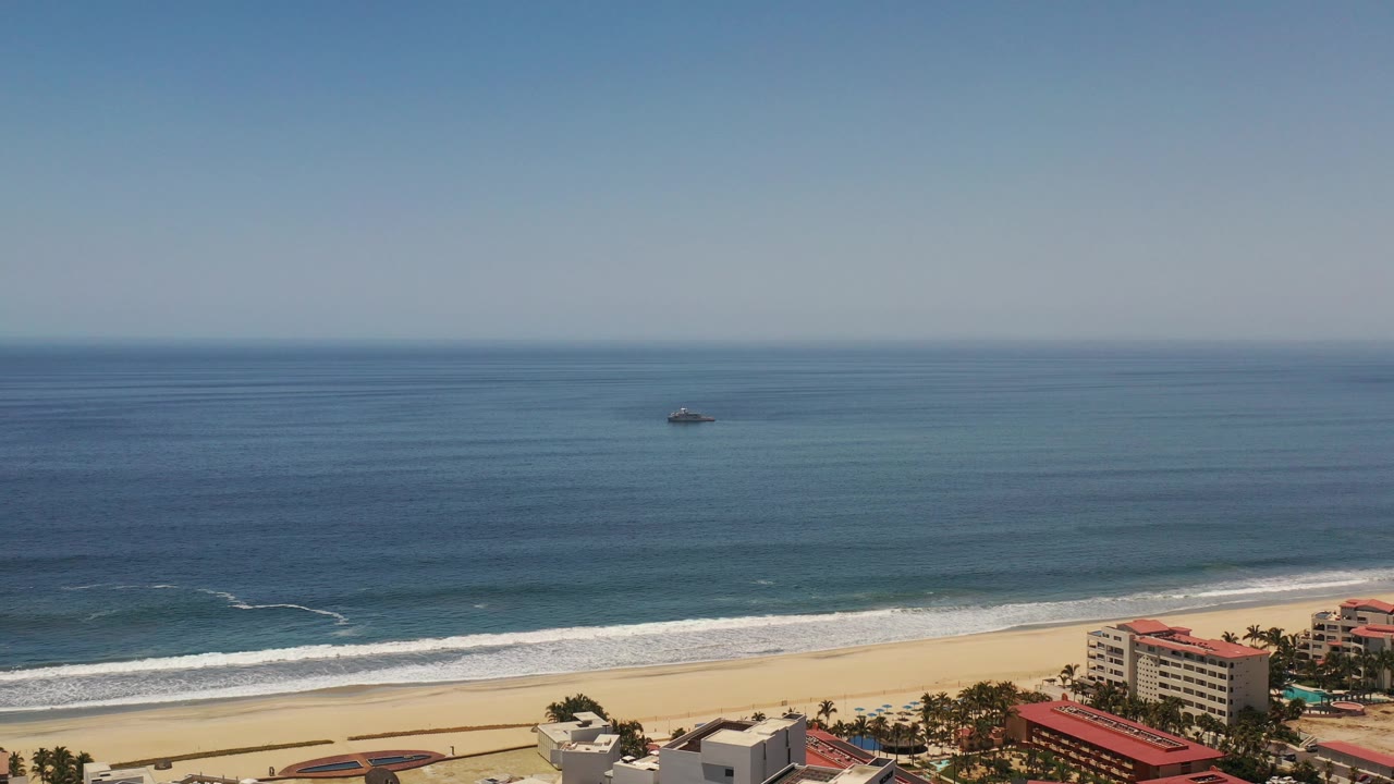 Small vessel on open water beyond surf and a sandy beach with coastal buildings.