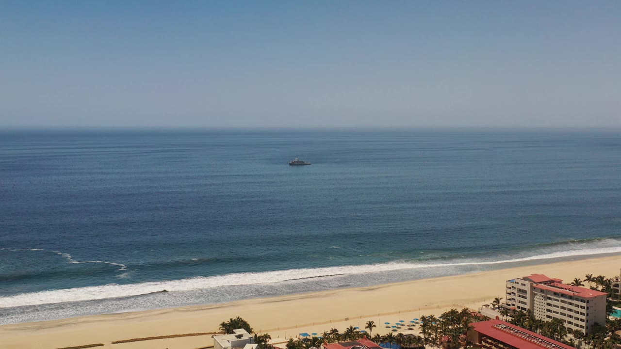 Los Cabos aerial-style view with the horizon high in frame, white breakers moving toward the sand, a small offshore boat, and no visible people.
