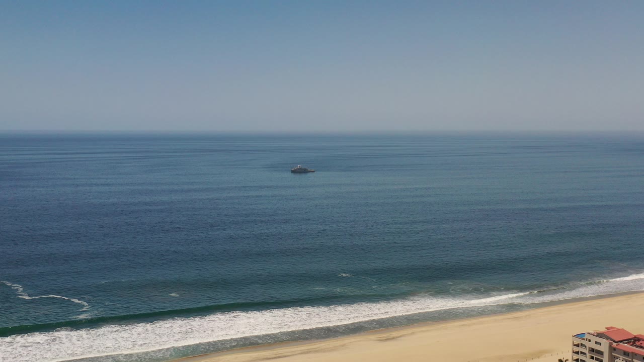 Opening frame of Los Cabos ocean water with white surf along the sandy beach, a small dark object offshore, a sliver of beachfront building at lower right, and no visible people.