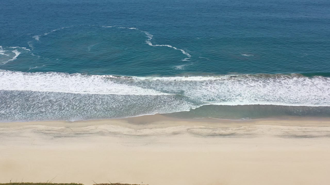 Aerial shoreline with blue ocean, white surf, and pale sand.