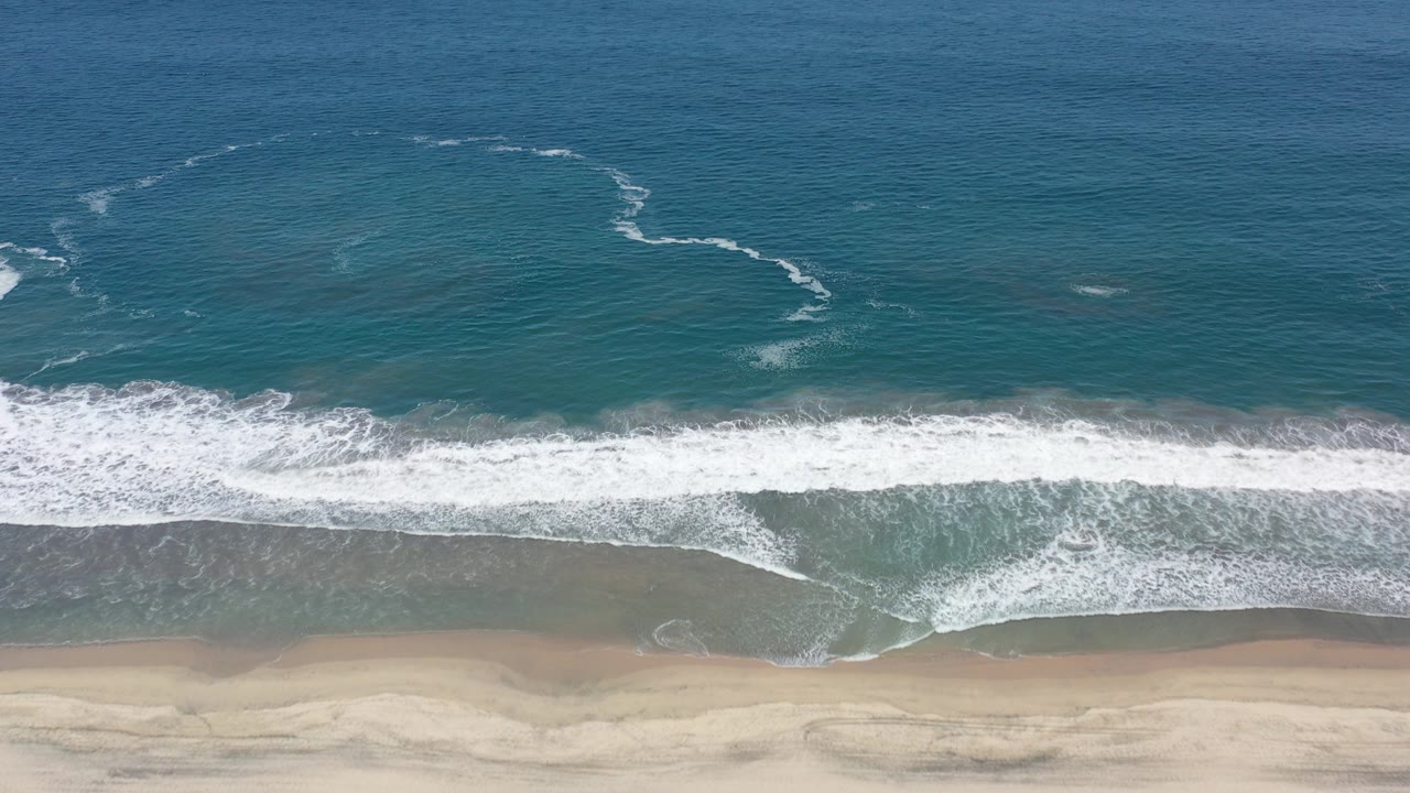 Overhead aerial frame from Los Cabos, Mexico showing teal-green water, a white surf line, wet sand, and no visible people at the start of the clip.