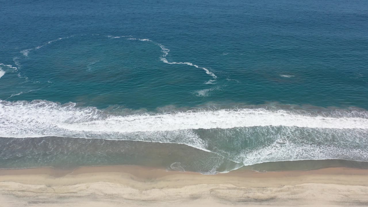 Overhead Los Cabos beach view with a broader white foam band crossing the frame and pale sand visible below, with no visible people.