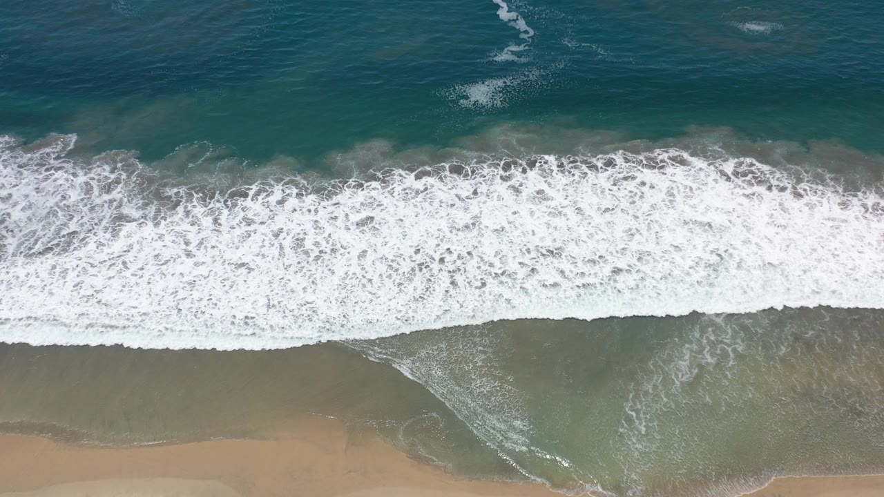 Top-down view of emerald ocean and white surf meeting tan sand in Los Cabos, Mexico, with no visible people.