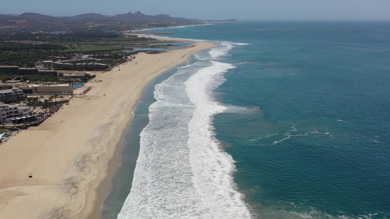 Aerial view of a sandy beach, breaking waves, ocean, beachside buildings, and distant hills.