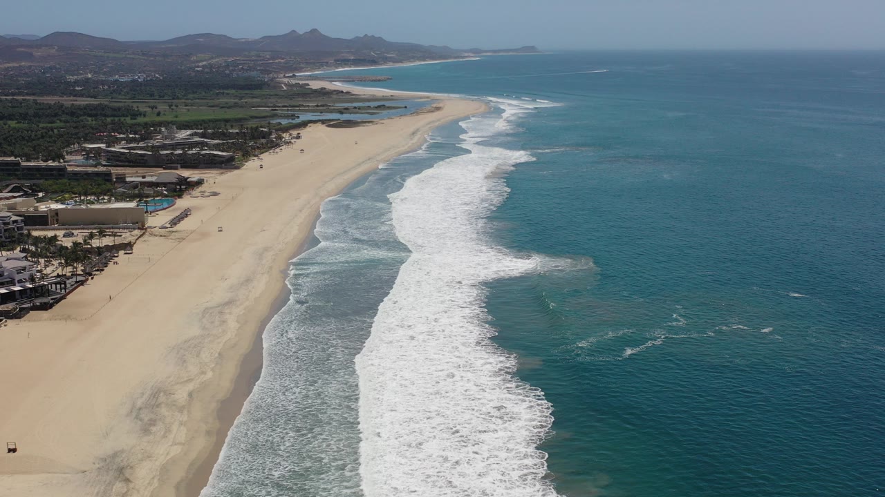 Aerial view of sandy beach, surf, ocean, coastal buildings, and distant hills.