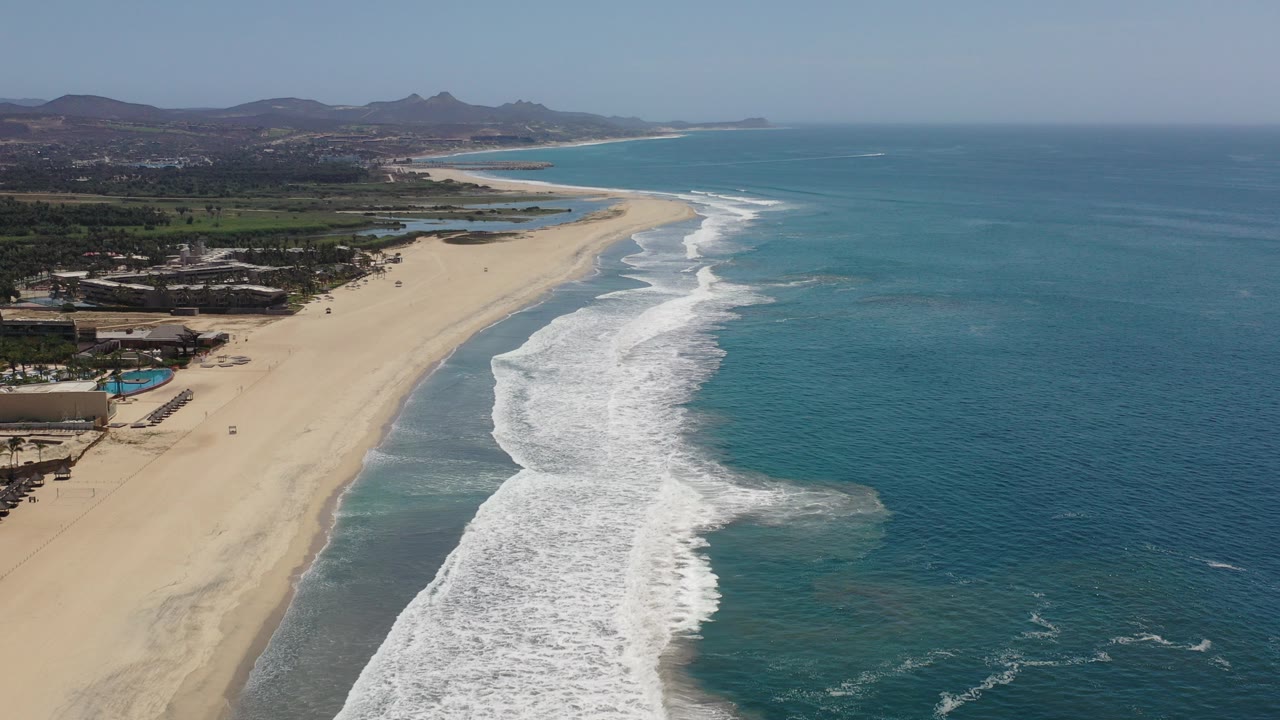 Aerial view over the Los Cabos coastline with a wide sandy beach, white surf bands, blue ocean to the right, low buildings at left, distant hills, and no visible people.