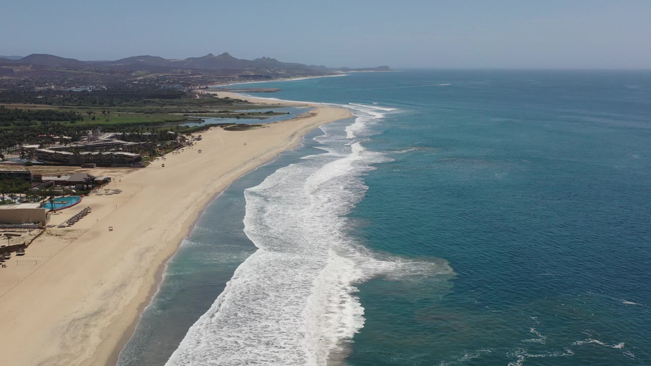 Coastal aerial with beach, waves, buildings, and hills.