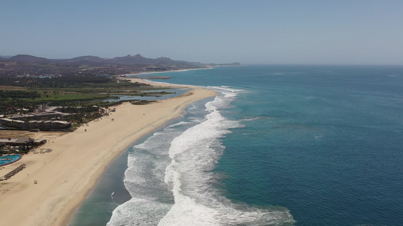 Aerial view over a Los Cabos beach at the start of the clip, with blue ocean on the right, white surf along the sand, inland greenery and hills, and no visible people.