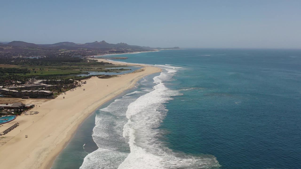High aerial frame over Los Cabos coastline, showing the long sand strip angling into the distance, stacked white wave lines, blue water, low inland terrain, and no visible people.