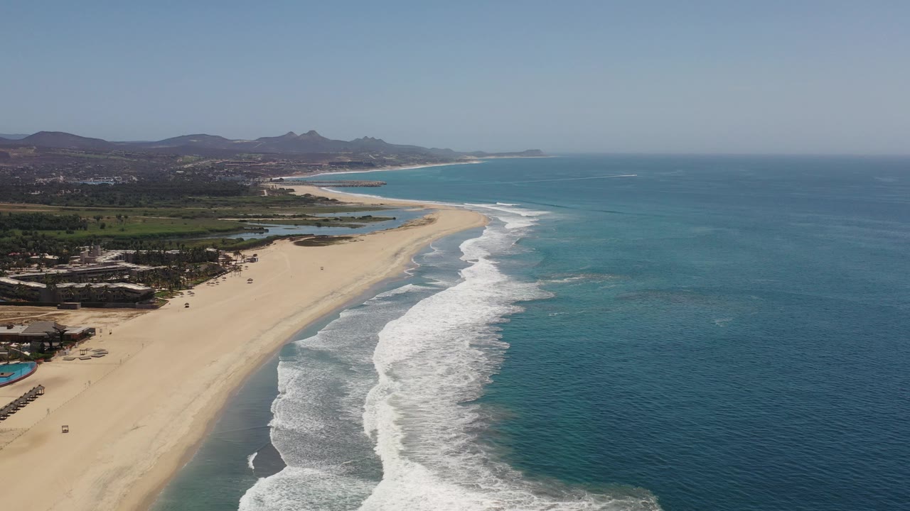 Drone view of surf washing onto a curved beach with hills inland.