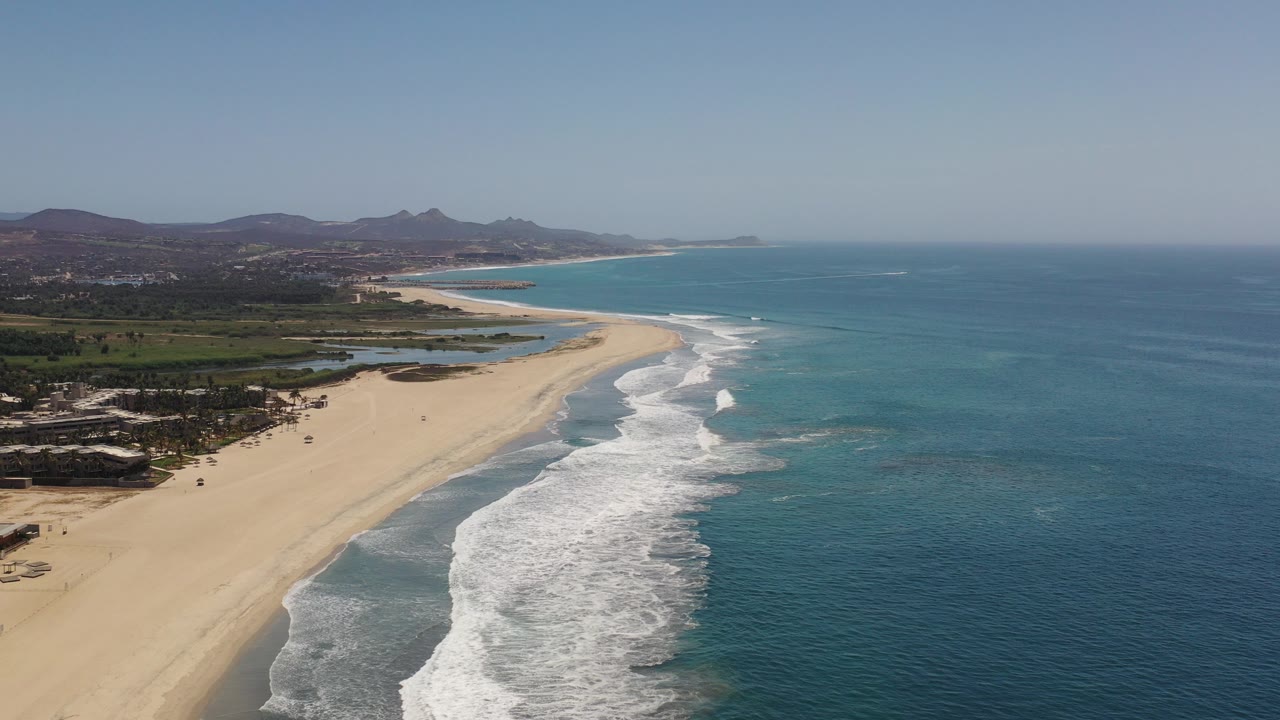 Aerial view of a sandy beach, white surf, blue ocean, green inland area, and distant hills.