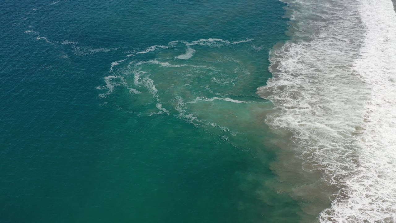 Los Cabos aerial water texture with curved foam marks over teal water and a dense surf strip on the right, with no visible people.
