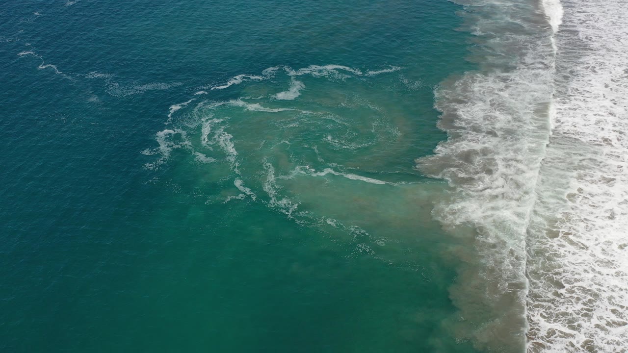 Overhead view of teal water and white surf foam washing along the right side of a Los Cabos beach, with no visible people.
