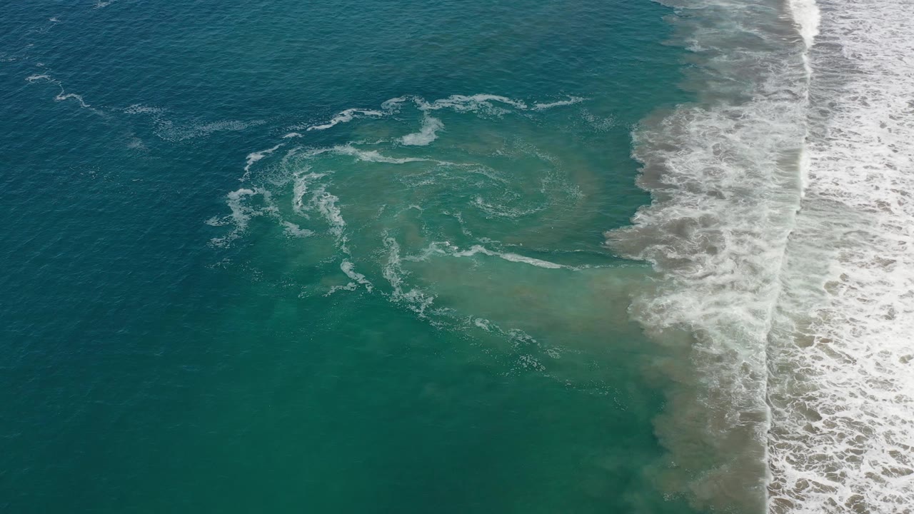 White foam forms curved streaks over green-blue Los Cabos water while a brighter band of surf breaks at the right, with no visible people.