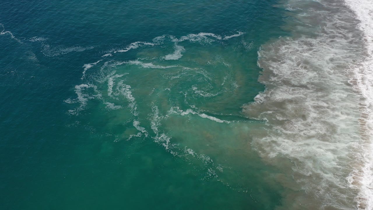 Overhead view of teal ocean water in Los Cabos, Mexico, with white surf on the right edge and a loose circular foam pattern near center, no visible people.