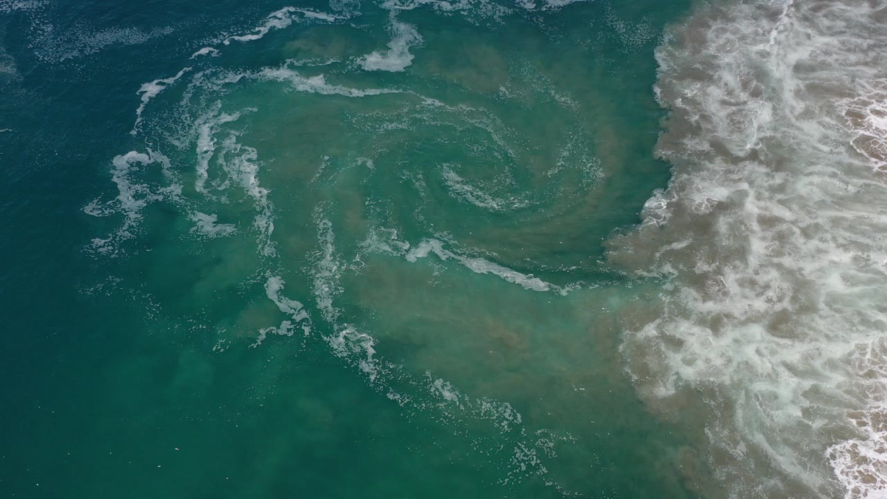 Overhead view in Los Cabos, Mexico of white surf foam beginning a circular shape in turquoise water, with breaking waves on the right and no visible people.