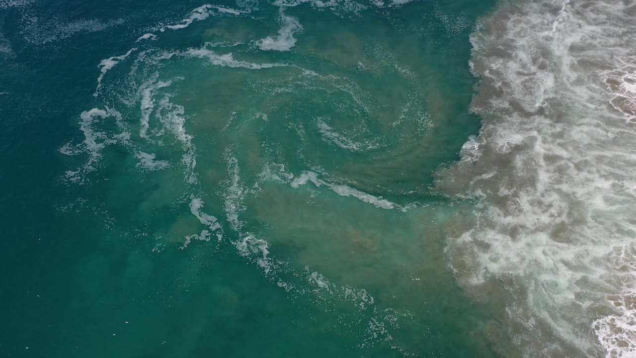 Overhead Los Cabos ocean frame with a clearer foam ring over green-blue shallows, darker water at left, breaking surf at right, and no visible people.