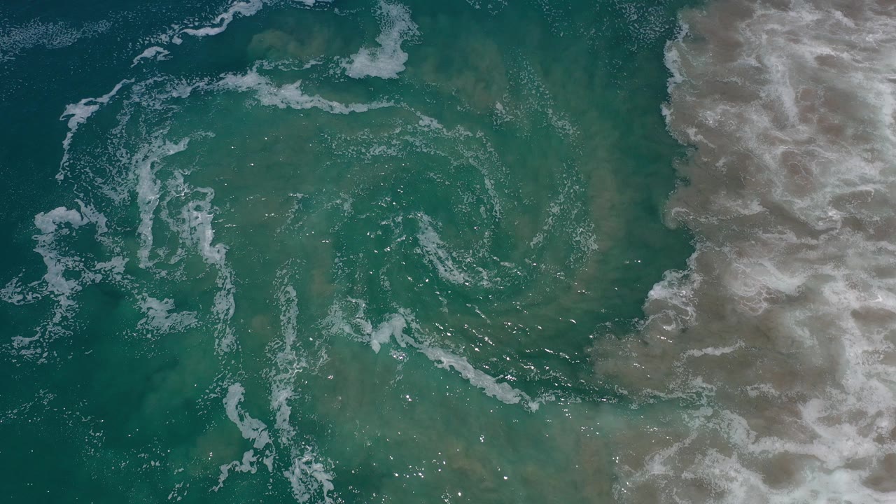Overhead view of turquoise ocean water in Los Cabos, Mexico, with circular white foam marks and a pale surf band along the right edge, no visible people.