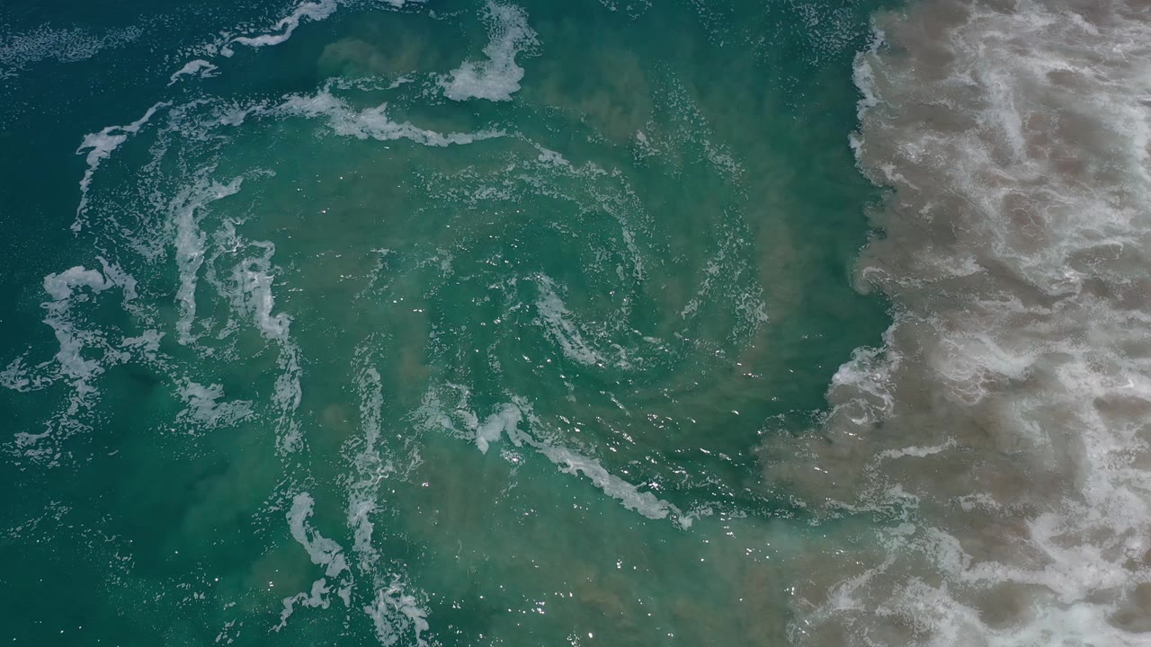 Green-blue ocean surface in Los Cabos, Mexico, with a loose ring of white foam near the center-left and breaking surf filling the right side, no visible people.