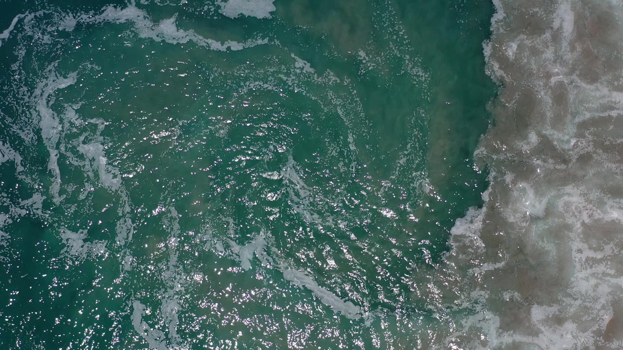 Overhead Los Cabos shoreline frame with foamy surf curling across turquoise water and a vertical strip of wet sand at right, with no visible people.