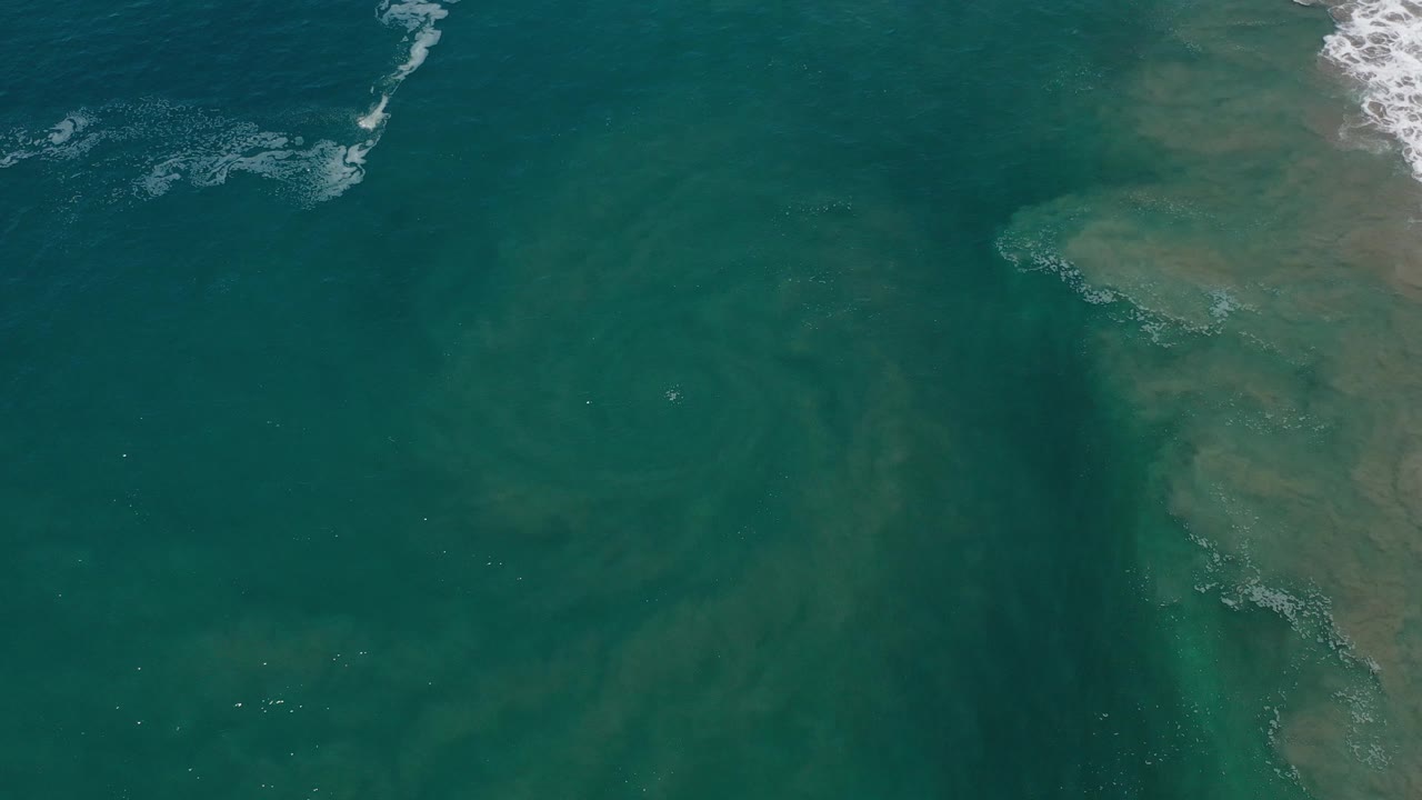 Overhead view of green-blue ocean water in Los Cabos, Mexico, with a dark swirling eddy and small white wave foam near the top and right edges, no visible people.