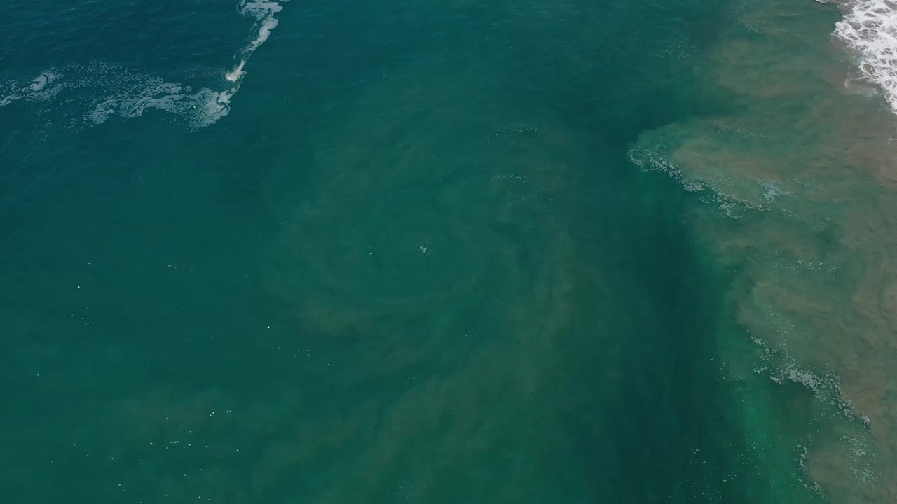 Overhead Los Cabos ocean frame with the eddy slightly left of center, pale green water gradients, and a thin strip of white foam entering from the upper edge, no visible people.