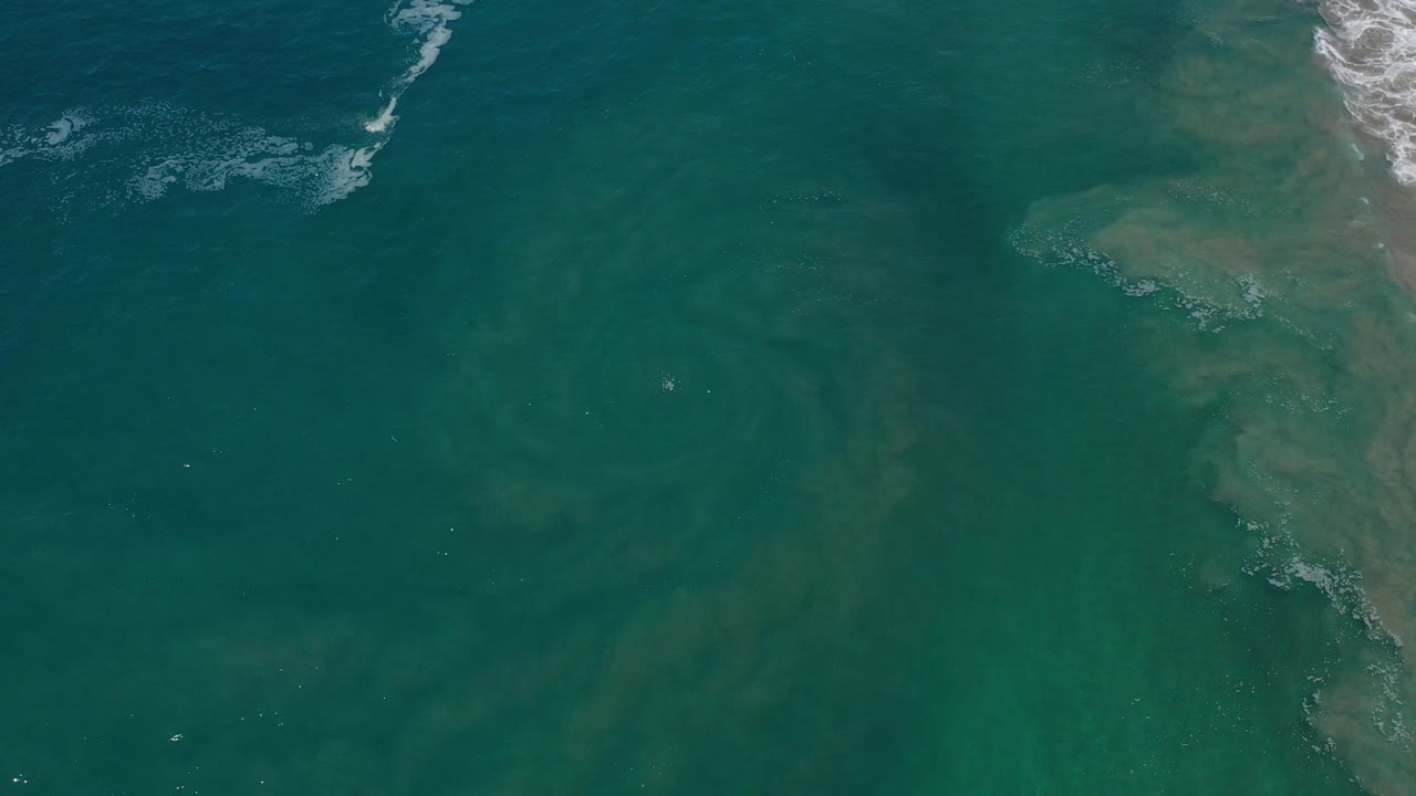 Overhead view of teal ocean water near the Los Cabos coastline, with white surf at the upper edge, rocky brown shore on the right, and no visible people.