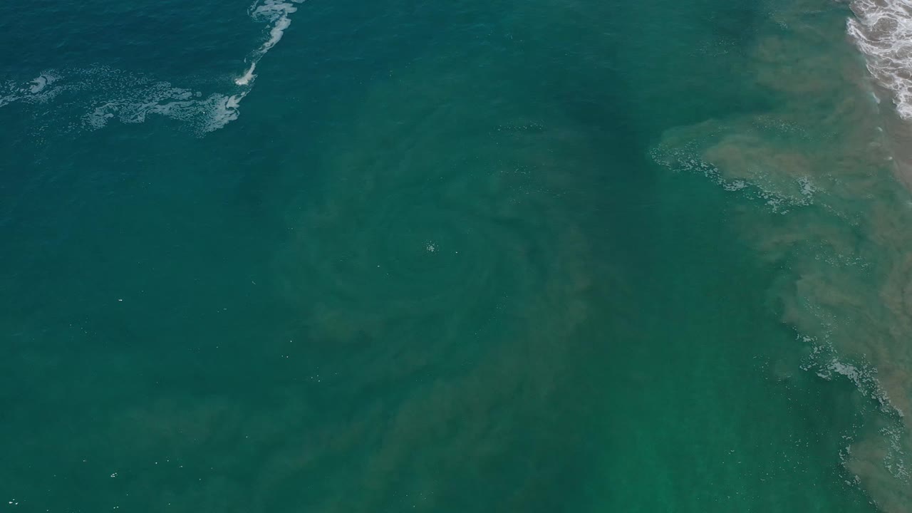 Aerial view over Los Cabos ocean water, with darker green patches across the surface, a strip of white surf near the top, and no visible people.