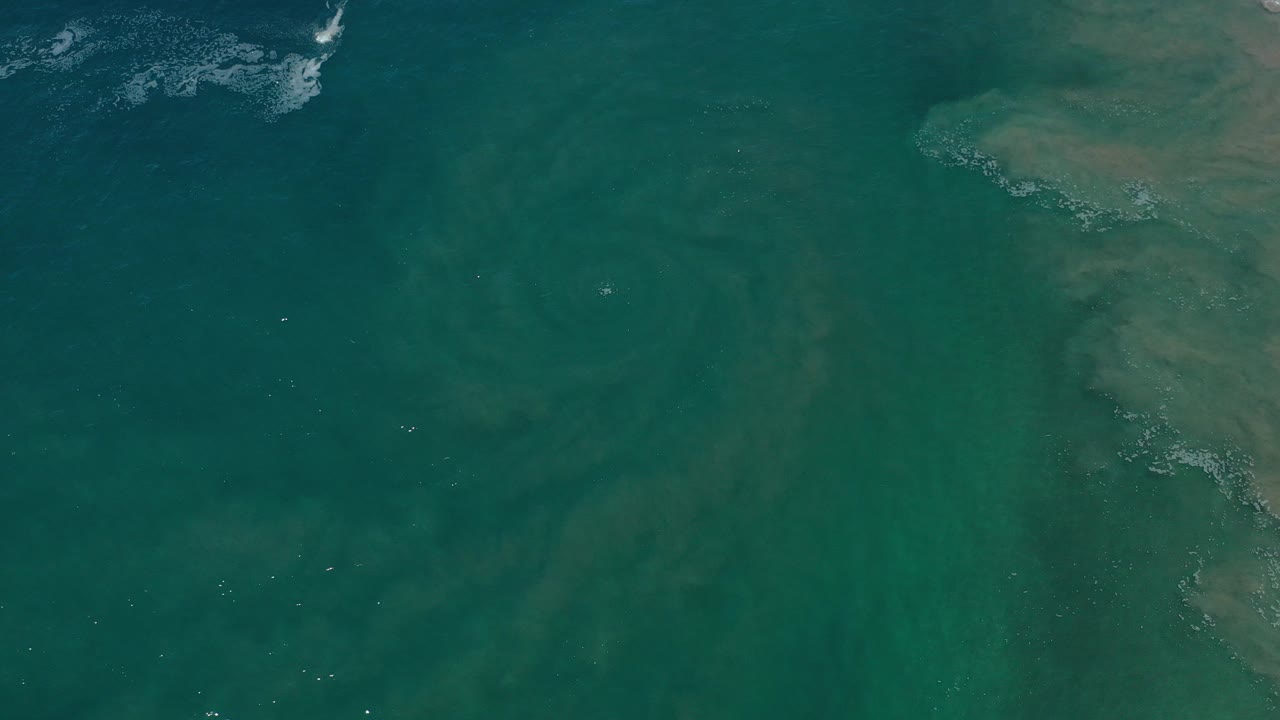 Opening overhead view near Los Cabos, Mexico, showing teal water, faint pale streaking, darker patches, a tan mass at the right edge, and no visible people.