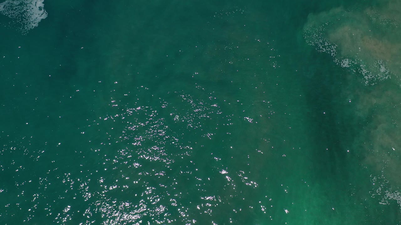 Top-down view of green ocean water in Los Cabos, Mexico, with bright sun glitter on ripples, faint pale patches under the surface, and no visible people.