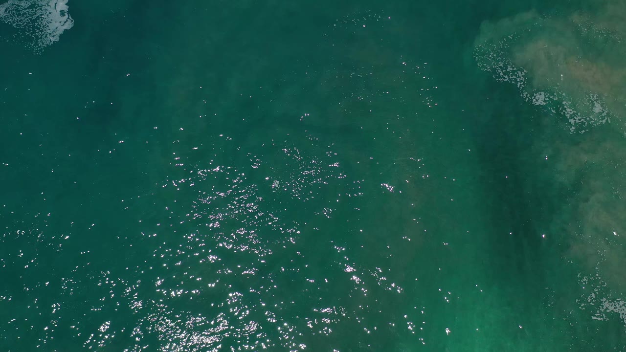 Aerial frame over Los Cabos green water, where small ripples catch light and a pale submerged sand or current patch sits near the upper right, with no visible people.
