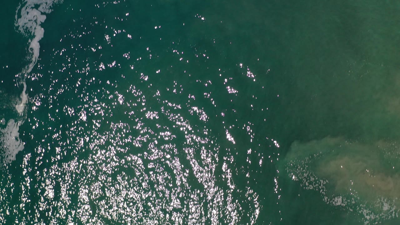 Overhead view of emerald ocean water in Los Cabos, Mexico, with bright sun reflections across the left half, a foam line along the left edge, and no visible people.