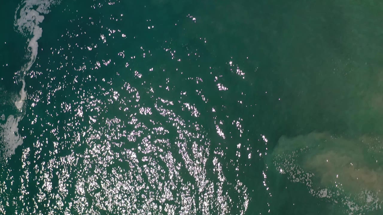 Aerial view of Los Cabos ocean water with concentrated sun glitter near the left center, white foam tracing the left edge, pale underwater sand on the right, and no visible people.