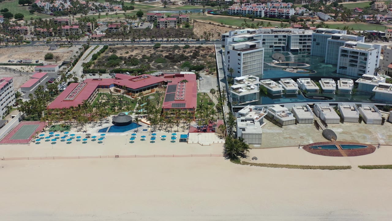 Aerial view of beachfront buildings, sand, palms, and umbrellas.