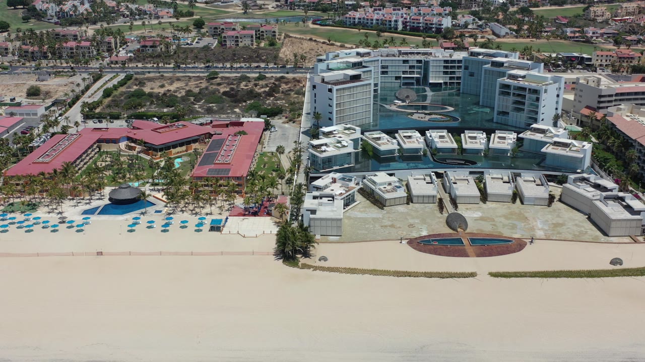 High aerial view in Los Cabos, Mexico, with a broad sandy beach in front of modern beachfront buildings, pool decks, palms, inland roads, and no visible people.