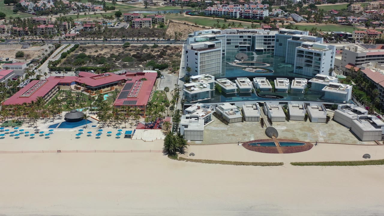 Aerial frame over Los Cabos beachfront development, showing a red-roofed building beside palms and blue pool water, with taller white buildings and no visible people.