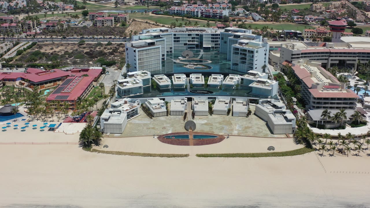 Opening frame of a large white beachfront hotel in Los Cabos, Mexico, with pale sand in front, blue pool areas to the left, inland buildings behind, and no visible people.